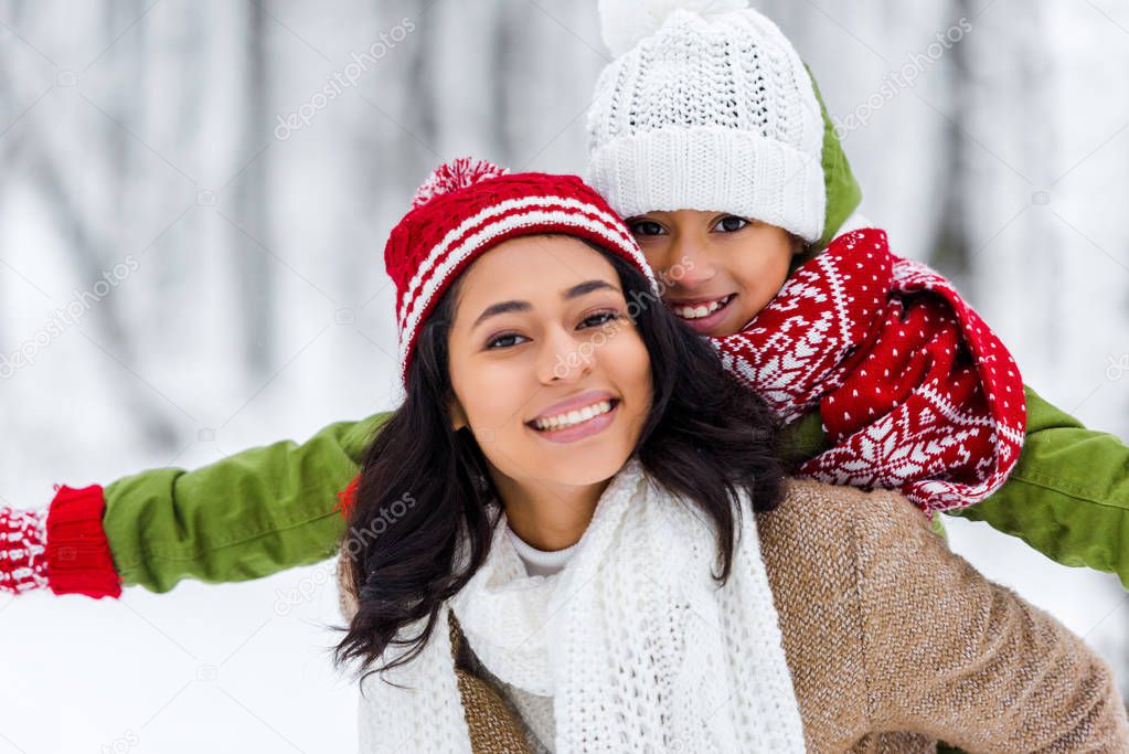 beautiful african american woman piggybacking cheerful daughter and looking at camera in winter park