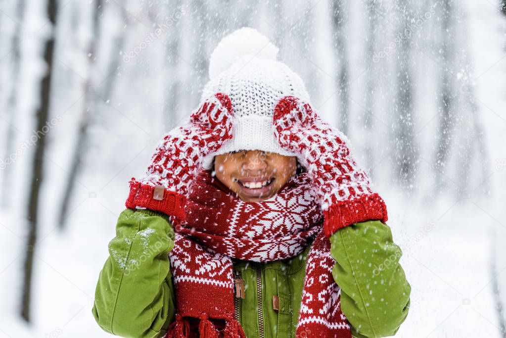 smiling african american child with knitted hat pulled over eyes during snowfall in winter park