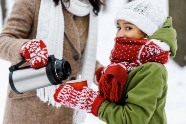 cropped view of woman in warm clothing pouring tea from thermos in cup holding by daughter looking at camera in winter clipart