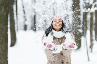 attractive african american woman throwing snow in park and smiling in winter forest clipart