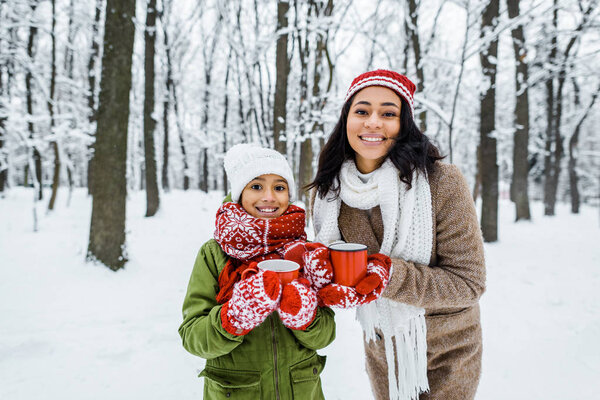 attractive african american mother and cute daughter holding cups of tea and smiling while looking at camera in snowy forest