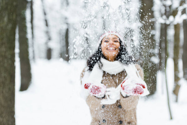 привлекательная американка из Африки бросает снег в парк и улыбается в зимнем лесу
