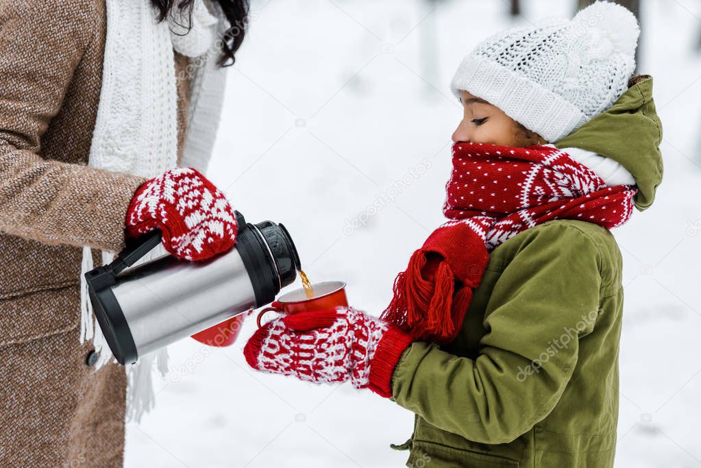 cropped view of mother pouring tea from thermos in red cup holding by african american daughter in winter