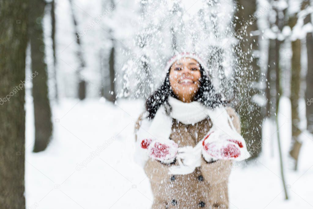 attractive african american woman throwing snow in park and smiling in winter forest