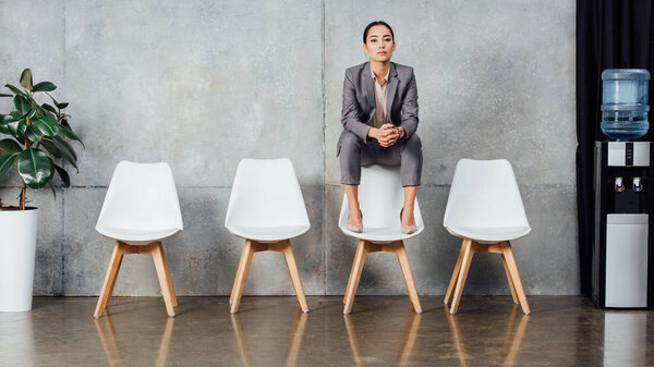 serious asian businesswoman in formal wear sitting on chair and looking at camera in waiting hall