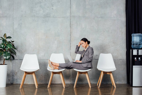 pensive asian businesswoman in formal wear sitting on chairs and using laptop in waiting hall