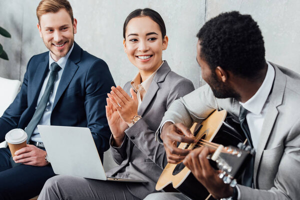 улыбающиеся мультикультурные бизнесмены сидят и слушают, в то время как африканский американец играет на гитаре в зале ожидания

