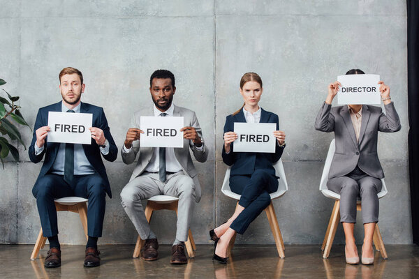 multiethnic businesspeople sitting and holding cards with 'fired' and 'director' words in waiting hall