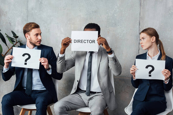 multiethnic businesspeople holding cards with 'director' word and question marks in waiting hall
