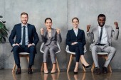 Lächelnde multiethnische Geschäftsleute sitzen auf Stühlen und jubeln mit geballten Fäusten in der Wartehalle
