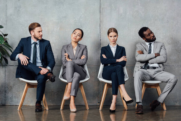 dissatisfied multiethnic businesspeople sitting on chairs with arms crossed in waiting hall