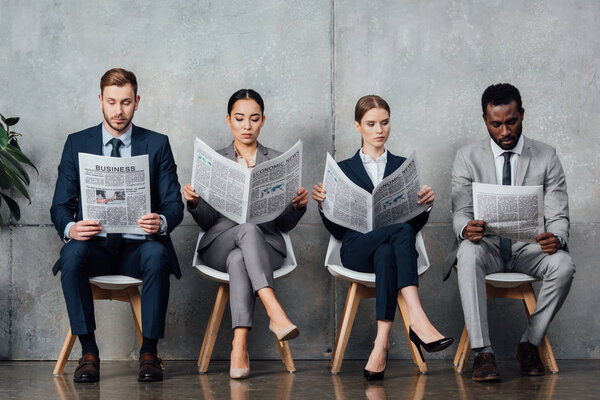 multiethnic businesspeople sitting on chairs and reading newspapers in waiting hall