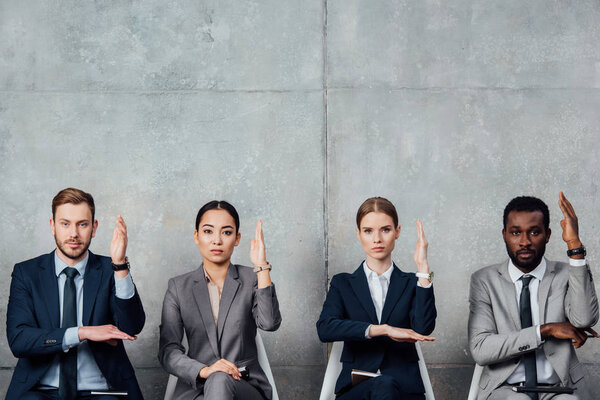 focused multiethnic businesspeople sitting on chairs with raised hands ready to answer in waiting hall