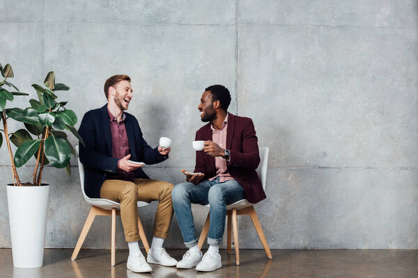 happy multiethnic men sitting, drinking coffee and talking in waiting hall