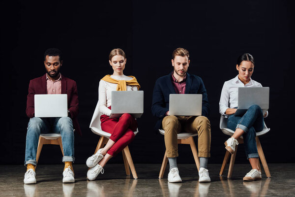 multiethnic group of casual businesspeople sitting on chairs and using laptops isolated on black