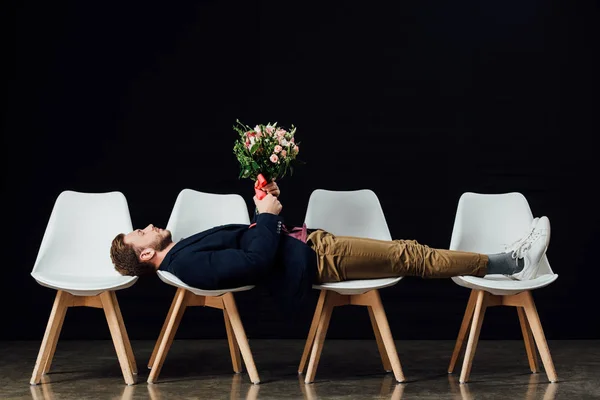 man with eyes closed lying on chairs and holding flowers isolated on black