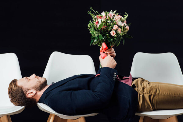 Красивый мужчина в повседневной одежде лежит на стульях и держит цветы изолированы на черном
