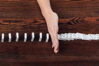 cropped view of woman preventing dominoes from falling on wooden desk clipart