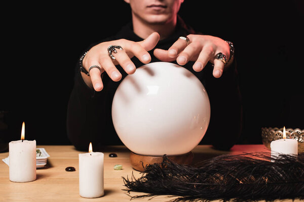 обрезанный вид человека, держащего руки над хрустальным шаром возле свечей, изолированных на черном
 
