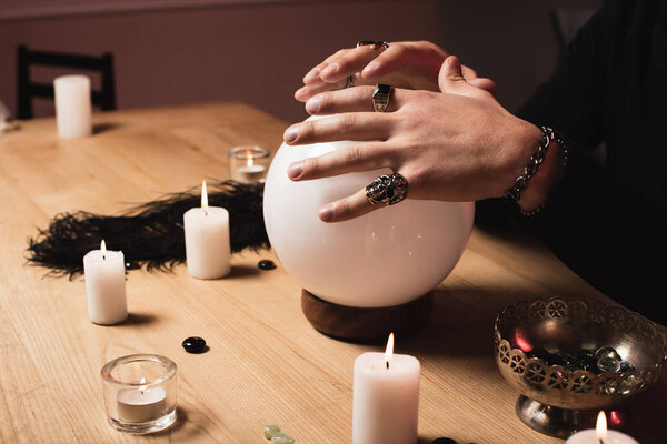 cropped view of psychic holding hands above magical crystal ball near candles 