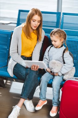  mother looking at digital tablet and sitting in airport with daughter  clipart
