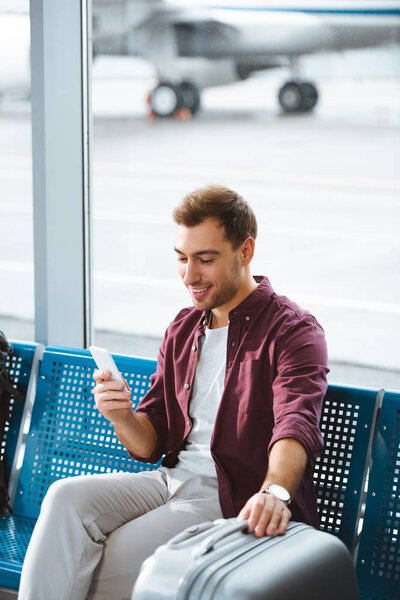 cheerful man looking at smartphone and smiling while holding luggage in airport 