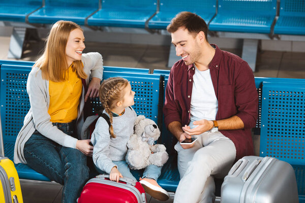 cheerful father smiling with daughter near suitcases and wife in departure lounge