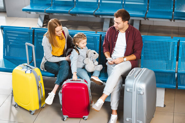 вид сверху на отца, смотрящего на дочь возле чемоданов и жену в салоне вылета
