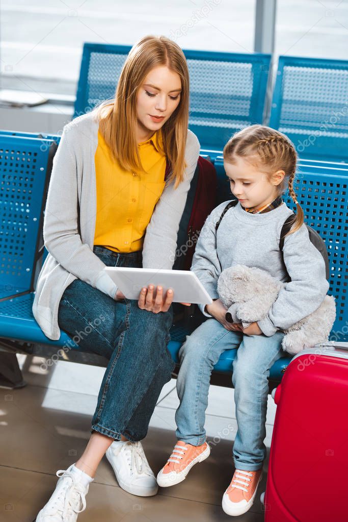  mother looking at digital tablet and sitting in airport with daughter 