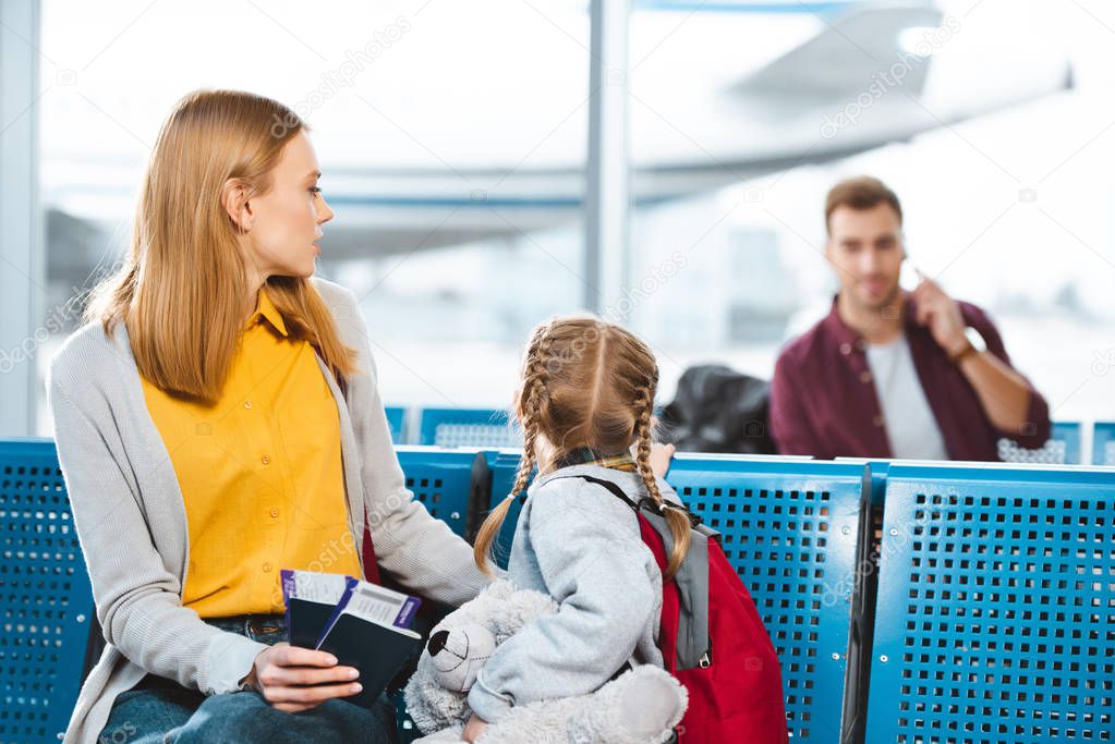 selective focus of mother holding passports with air tickets near daughter and looking at man in airport 