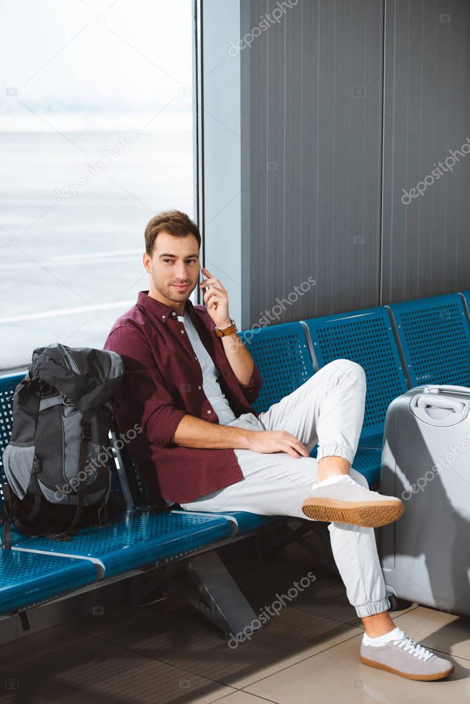 smiling man sitting in departure lounge and talking on smartphone 