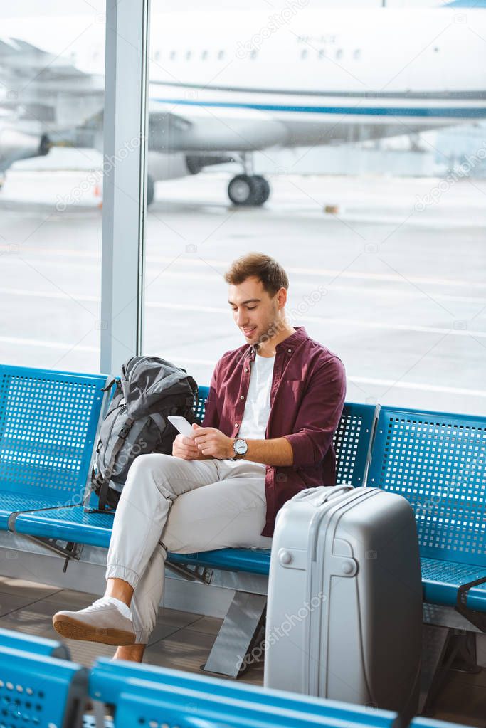 smiling man using smartphone while sitting in waiting hall in airport
