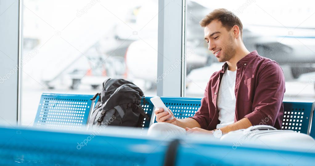 cheerful man holding smartphone and smiling while waiting in waiting hall