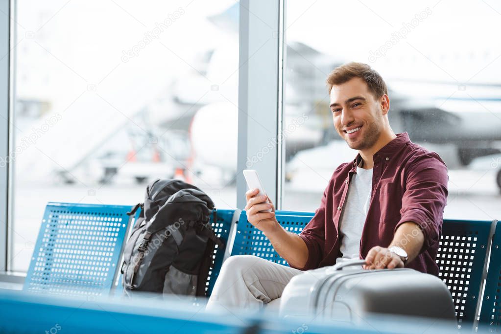 cheerful man looking at camera while holding smartphone and luggage in departure lounge