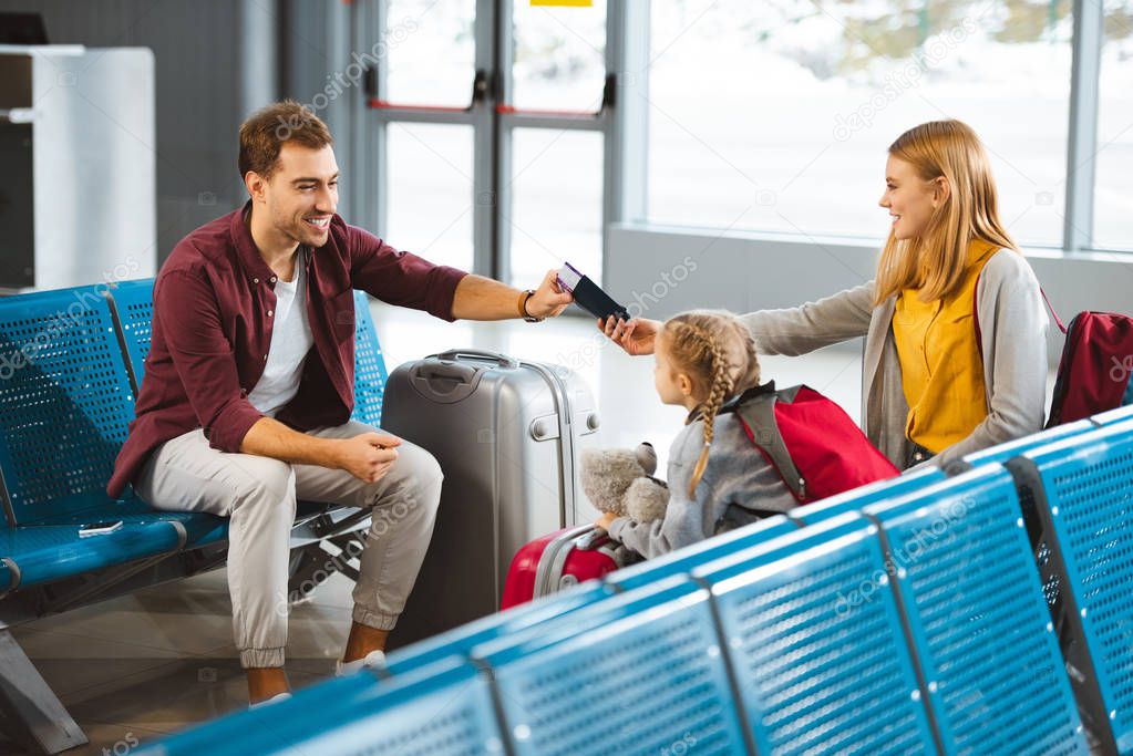 happy wife sitting with daughter and  giving passports with air tickets to husband in airport 