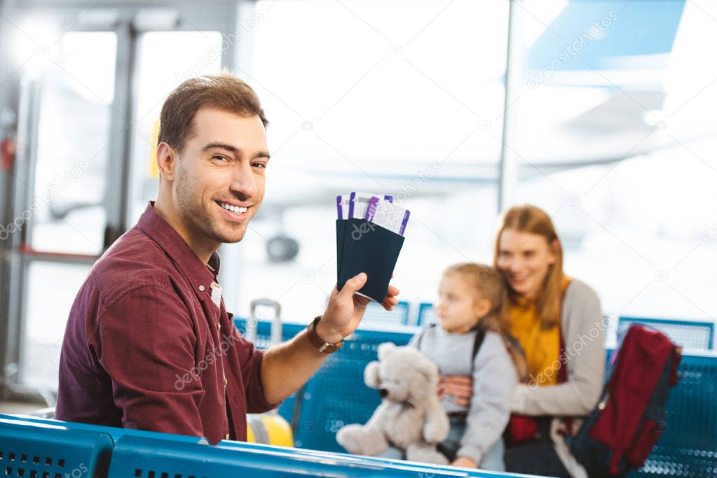 selective focus of handsome man smiling while holding passports with air tickets with wife and daughter on background 