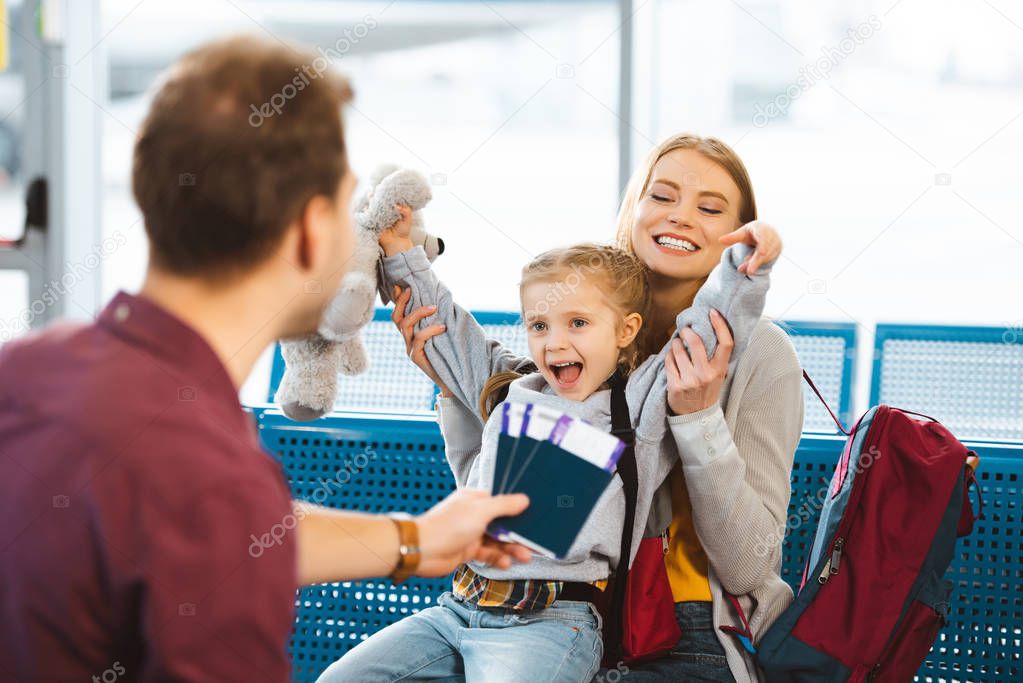 selective focus of cheerful daughter holding hands above head and smiling with mother near father with passports with tickets 