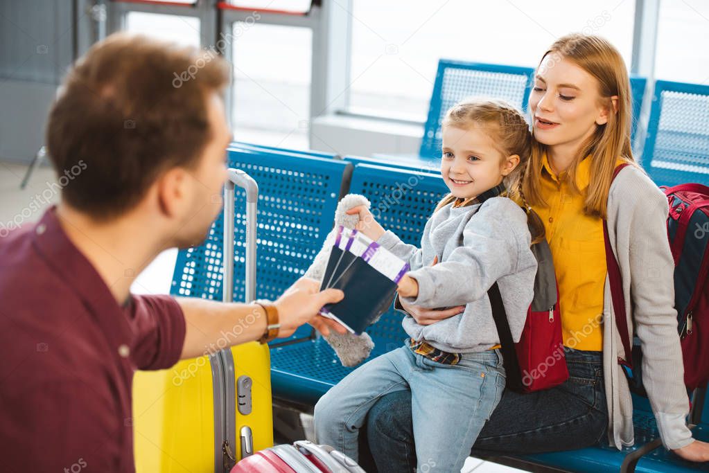 selective focus of cute daughter looking at dad with passports and air tickets while sitting with mom in departure lounge