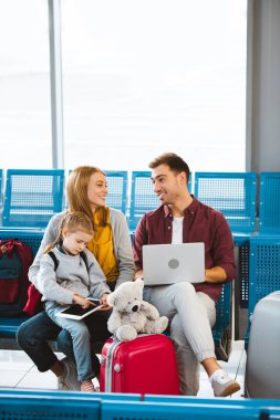 happy wife and husband looking at each other near daughter using digital tablet in waiting hall clipart