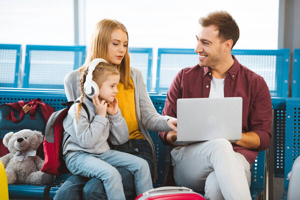 attractive woman talking to husband with laptop near daughter in headphones in airport 