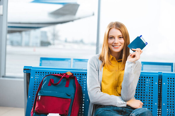 smiling woman holding passport with air ticket in airport near backpack