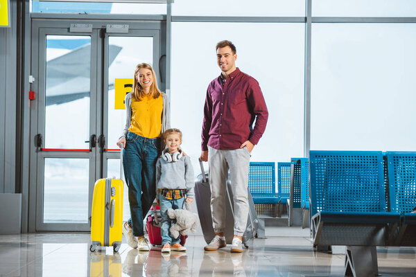 cheerful kid holding teddy bear and standing with mom and dad in departure lounge