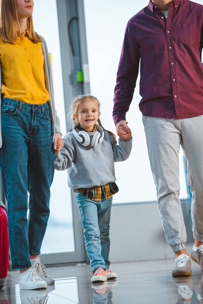 cheerful kid holding hands of father and mother in airport 
