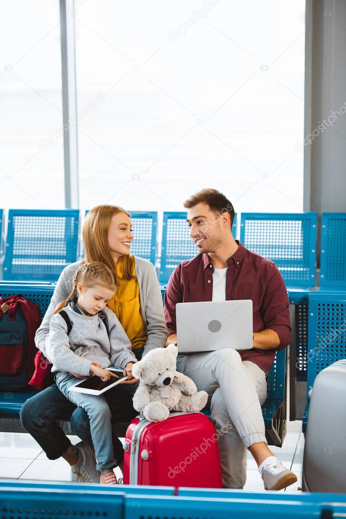 happy wife and husband looking at each other near daughter using digital tablet in waiting hall