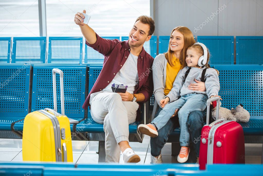 cheerful family taking selfie and smiling in airport near luggage