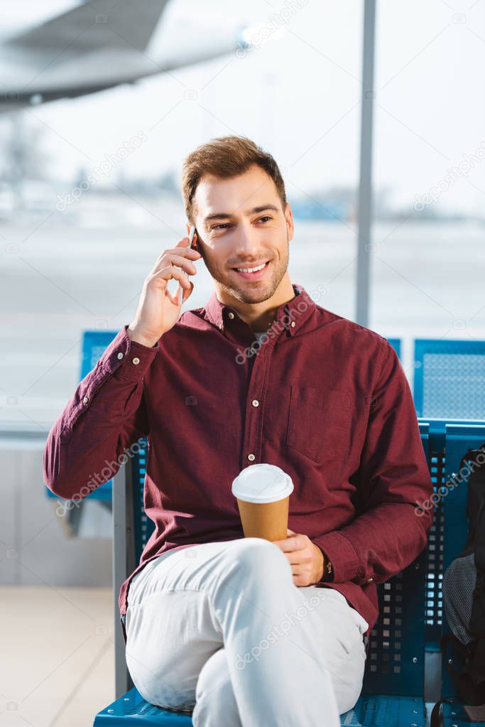 handsome man talking on smartphone and holding paper cup in hand in airport 