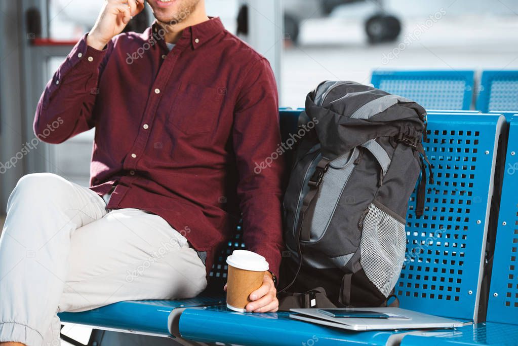 cropped view of man holding paper cup near backpack and laptop 