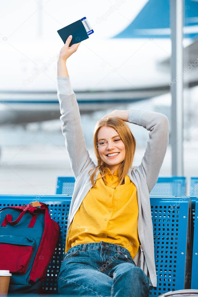 smiling woman holding passport above head in airport near backpack