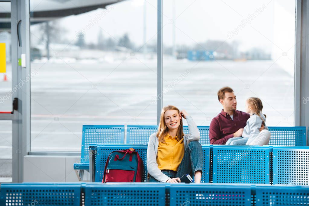 selective focus of smiling woman sitting in airport near backpack with people on background 