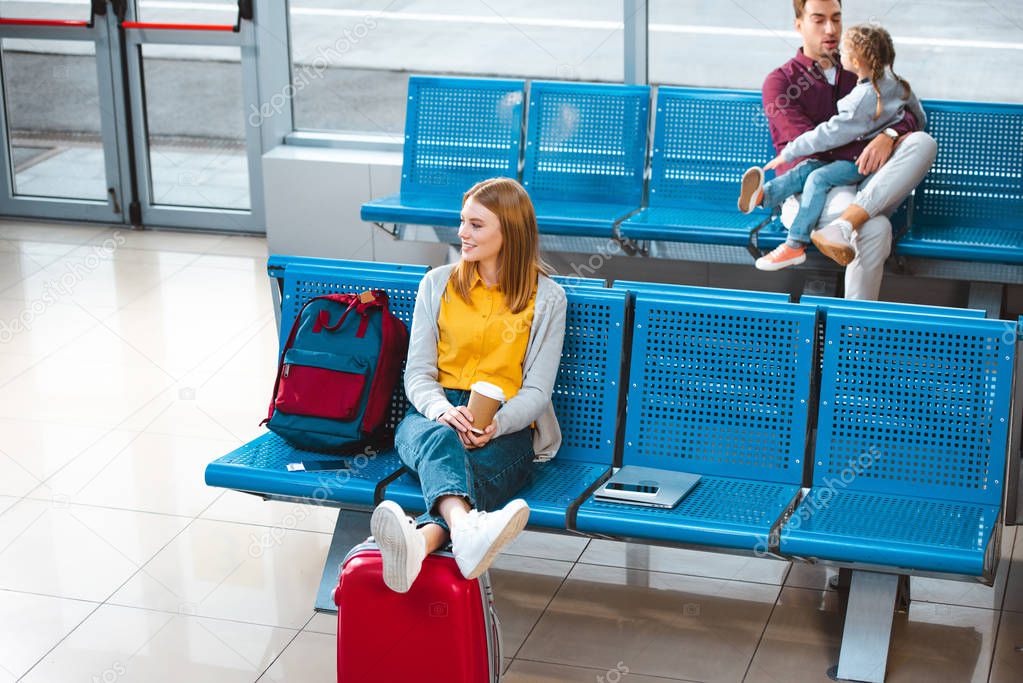 selective focus of smiling woman holding paper cup in airport near backpack with people on background 